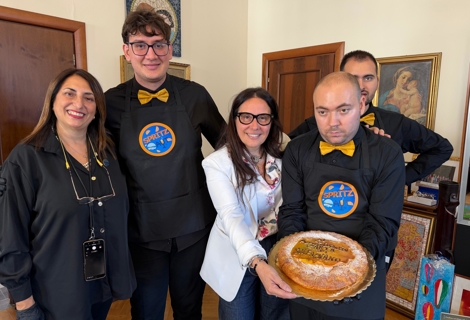 Il Ministro per le Disabilità Alessandra Locatelli posa insieme ai ragazzi di Spritz Aut, che indossano grembiuli neri con il logo del progetto. In primo piano viene mostrata una torta celebrativa dedicata all’anniversario della Carta di Solfagnano. La foto è scattata in un ambiente interno con quadri e opere decorative sullo sfondo.