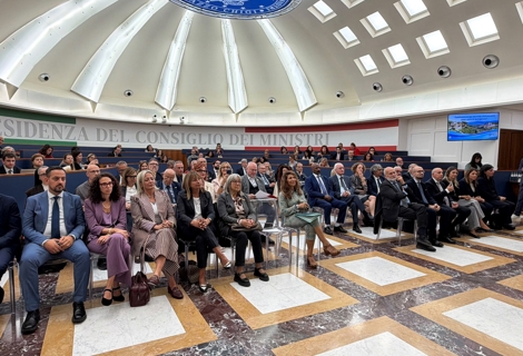Sala conferenze della PCM. L’ambiente è caratterizzato da una grande cupola illuminata da lucernari e dal logo della Presidenza al centro del soffitto. Sullo sfondo è visibile la scritta “Presidenza del Consiglio dei Ministri”, mentre un monitor mostra una slide dedicata alla celebrazione del secondo anniversario della Carta di Solfagnano. I partecipanti occupano diverse file di sedute in una disposizione ad anfiteatro.